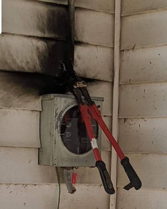 Ett elskåp med märken av brand och sot på väggen, en stor bultsax är placerad ovanpå skåpet.