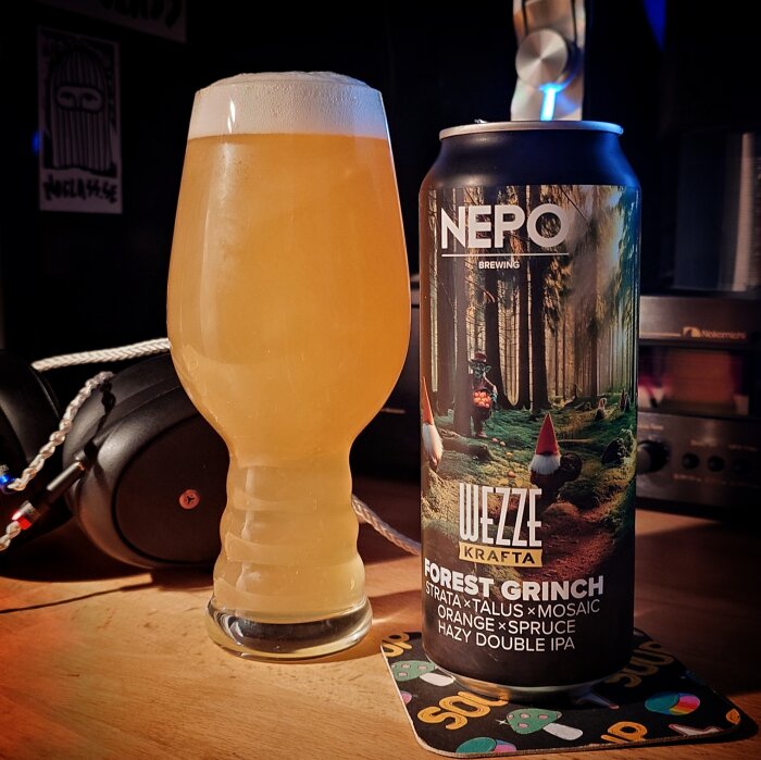 En burk NEPO Brewing Forest Grinch Hazy Double IPA står bredvid ett glas fyllt med samma grumliga öl, placerade på ett bord med hörlurar i bakgrunden.