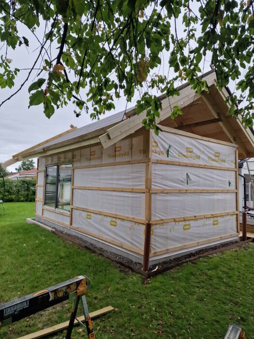 Hus under byggnation med vindduk och fönster/skjutdörr installerade, omgiven av grön gräsmatta och träd.