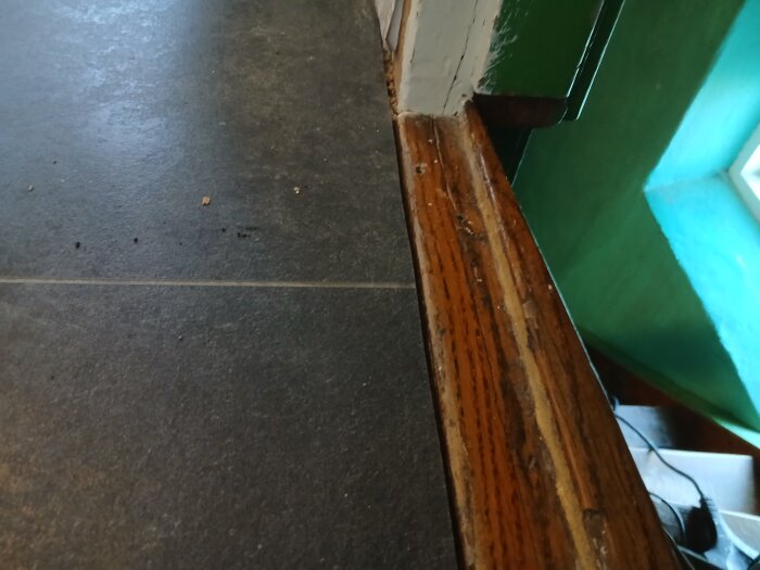 Tröskel i mörkt trä mot mörkt golv med en synlig glipa emellan, omgiven av gröna väggar och fönsterljus i bakgrunden.