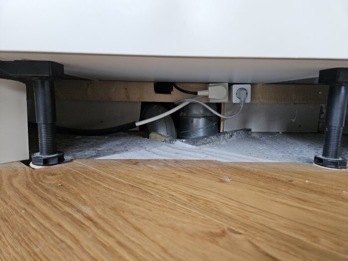 Undersida av köksskåp med synliga ventilationsrör, kablar och eluttag, samt parkettgolv med borttagna golvsocklar.