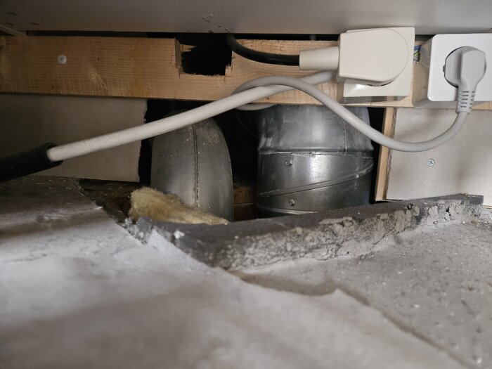 Ventilationsrör och elkablar synliga bakom golvsockel i ett kök, med isolering liggandes runt rören.