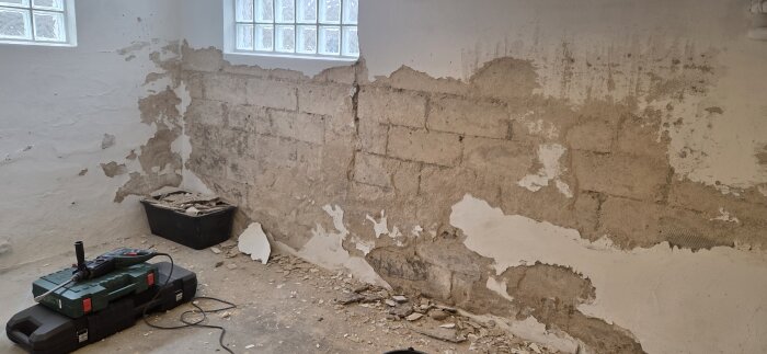 Vägg med delvis borttagen puts i källare, hålrum och sprickor syns. Verktygslåda och redskap står på golvet, murbruk är avlägsnat.