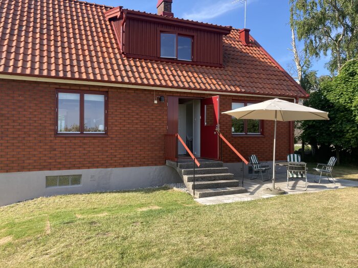 Tegelfritidshus i 70-talsstil med rött tak och entrétrappa, uteplats med parasoll och stolar i trädgården.