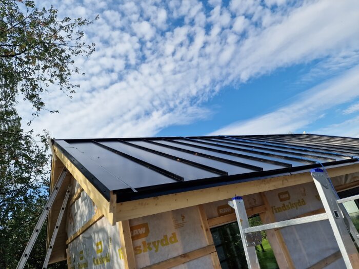 Plåttak monterat på byggnad med 32° taklutning, omgiven av en stege och träd, med blå himmel i bakgrunden.