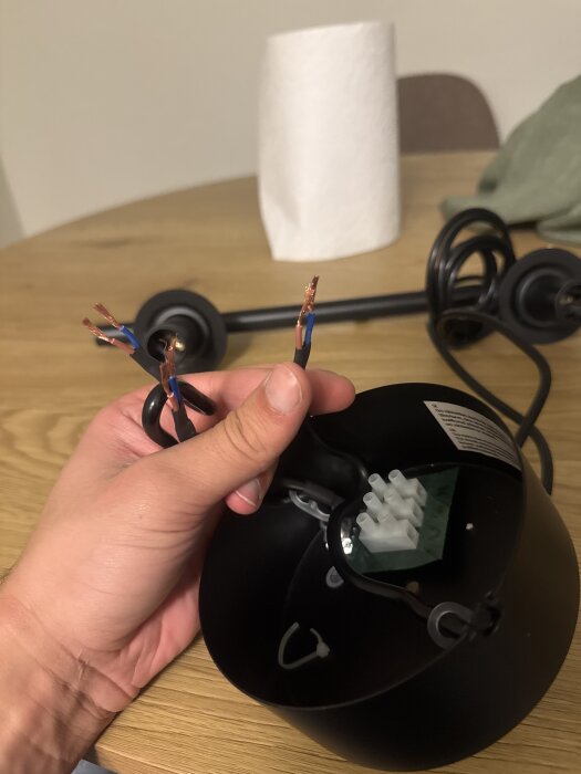 En hand håller upp tre elkablar, kopplingsdosan visas i bakgrunden på ett träbord.