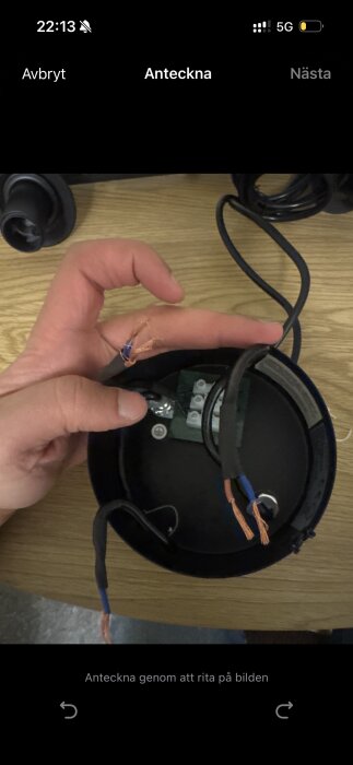 En hand håller elektriska trådar bredvid en kopplingsbox på ett bord. Fristående koppartrådar syns nära anslutningspunkten.