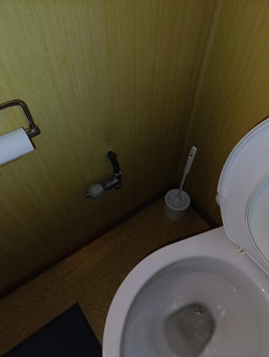 Toalett med borttagen vattenradiator, synlig röranslutning på väggen, gult tapet och golv samt toalettborste bredvid toalettstolen.