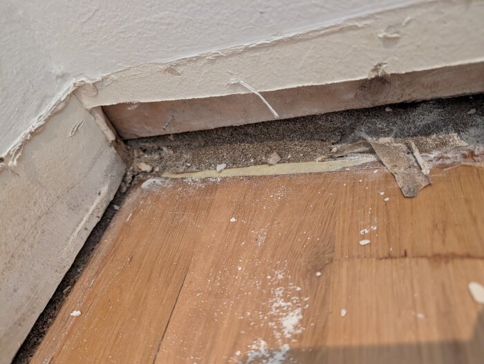 Nedre del av en vägg med borttagna lister, mörk parkett och sand vid kanten, troligtvis orsakat av vatten/dålig tätning.