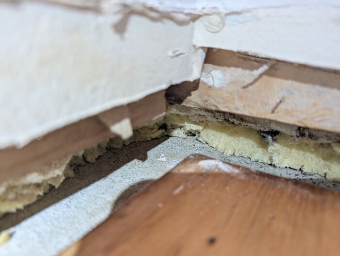 Närbild av ett parkettgolv där listerna tagits bort, visar isolering och fogskum runt betongväggarna. Tecken på mögel och smuts syns.