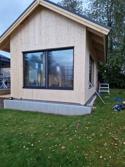 Ett nybyggt hus med stående panel av norrländskt trä och monterade hängrännor, omgiven av grön gräsmatta och träd i bakgrunden.