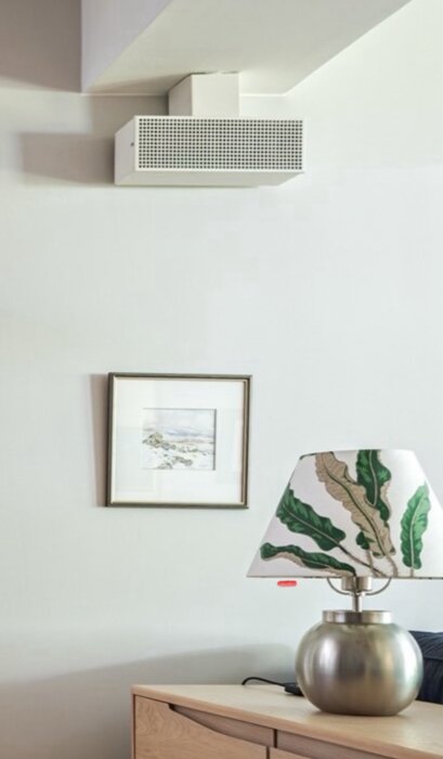 Väggmonterat vitt tilluftsdon ovanför en tavla och lampa på ett träbord i ett rum.