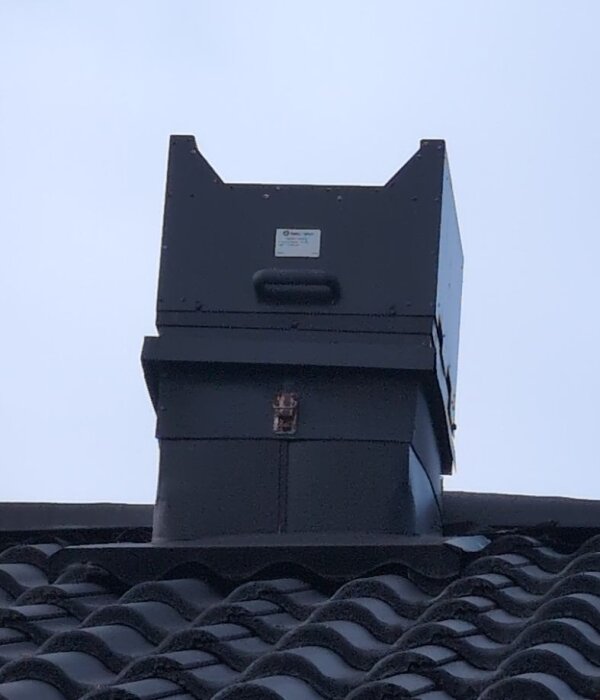 Svart takhuv eller ventilationskåpa på ett tak, fotograferad från marken med omgivande mörkgrå takpannor.