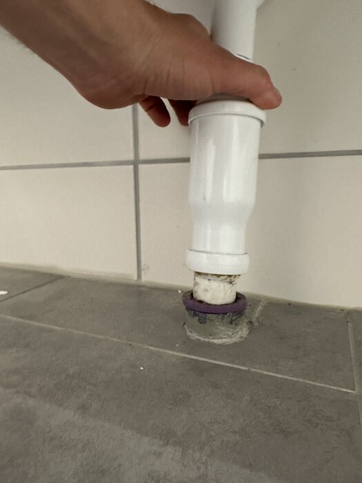 En hand håller i ett vitt avloppsrör som är instoppat i ett grått rör i golvet, med en lila muff synlig mellan rören.