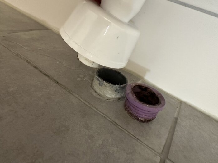 Två rör med olika dimensioner, ett vitt och ett grått, samt en lila muff visas vid ett avlopp under ett tvättställ.