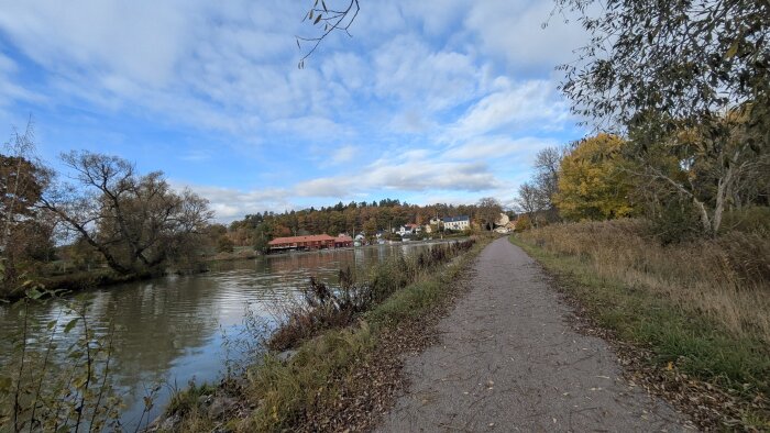 Gångstig längs en flod med träd och buskar i höstfärger, hus på andra sidan vattnet och en blå himmel med moln.