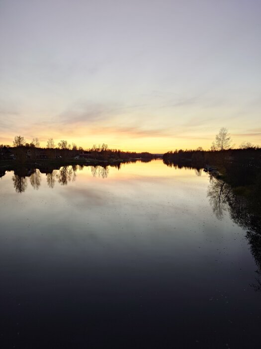 Solnedgång över en stilla flod, med träd på båda sidor och en klar himmel som reflekteras i vattnet.