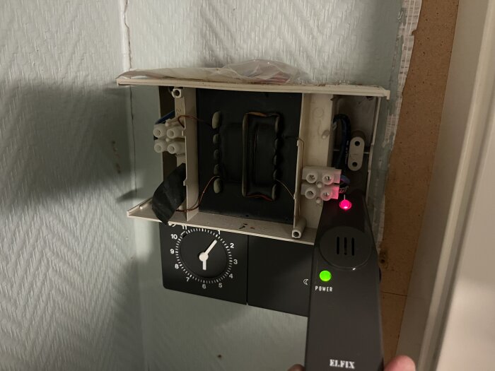 Strömdetektor som visar utslag på en brun kabel i en öppnad elbox med sladdkopplingar och en klocka, på väggen.