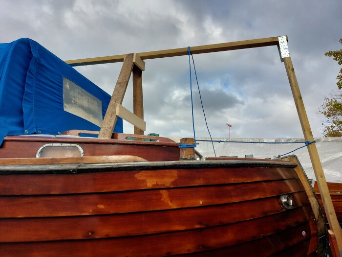 Träbåt med blå täckning och nybyggd täckställning av träreglar, markerade av byggbeslag, under en molnig himmel.