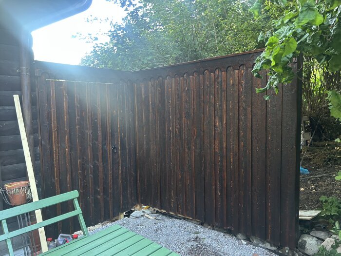 Brun träport i trädgård omgiven av grönska, sedd från grusgång med ett grönt träbord i förgrunden.