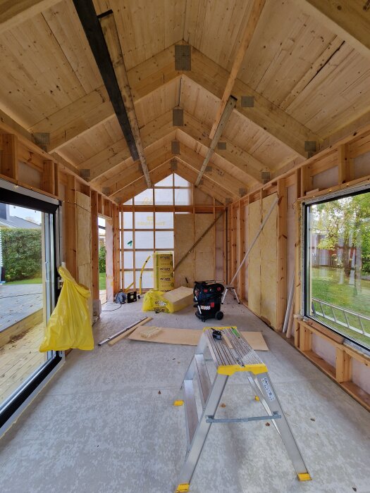 Interiör av ett byggprojekt med oisolerade väggar, en stege, isoleringsmaterial och arbetsverktyg på golvet.