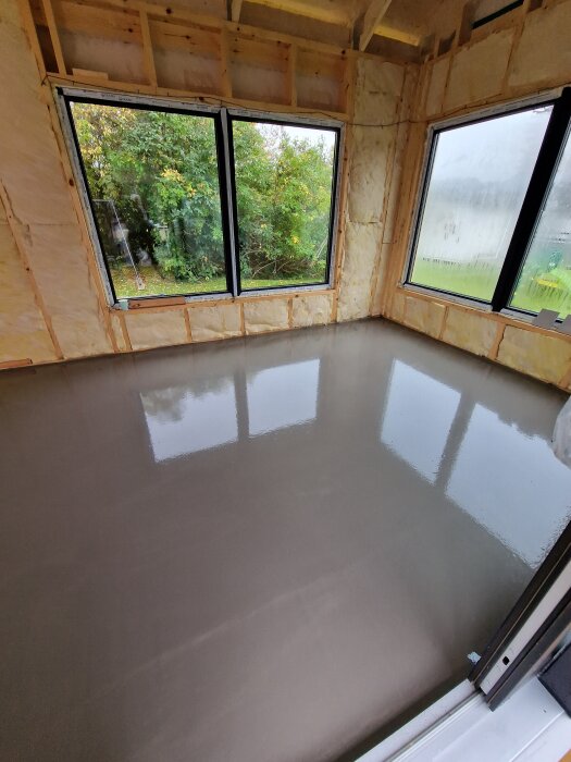 Rum under renovering med isolerade väggar och nylagt flytspackel på golvet, stora fönster vetter mot grönskande trädgård.