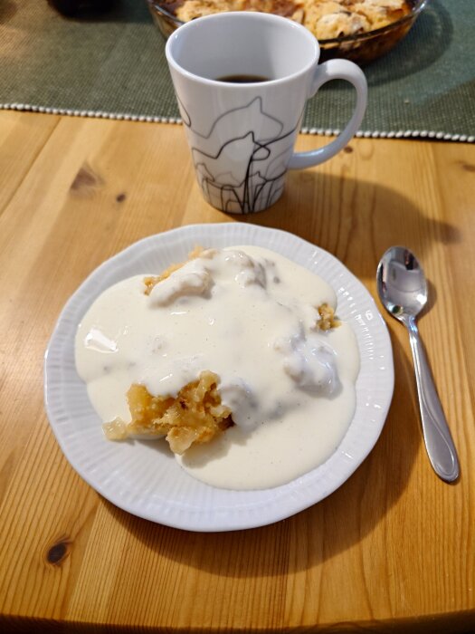 Äppelpaj med vaniljsås på tallrik, kaffekopp bredvid; på träbord.
