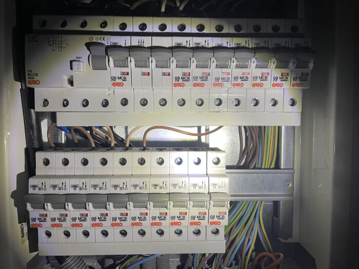 En elcentral med automatsäkringar och många kopplade kablar i olika färger, belyst av ficklampor.