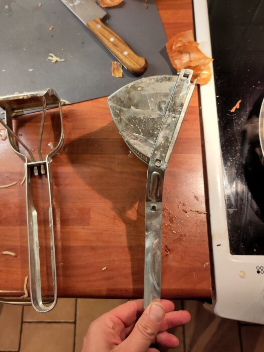 Metallpotatispress sönder efter användning, ligger på köksbänk med kniv och potatisskal runtom.