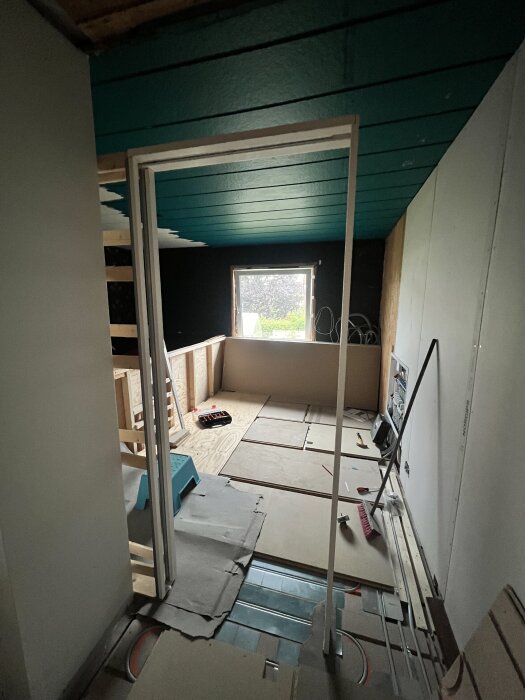 En byggarbetsplats med en dörrkarm monterad framför en vägg, omgivande verktyg och byggmaterial, och takpaneler i teal-färg.