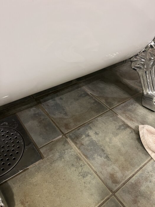 Klinkergolv med synlig fog och golvbrunn bredvid ett badkar på lejontassar, en fuktfläck märks nära brunnen.