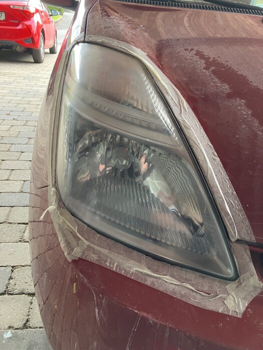 Närbild av en röd Prius-strålkastare under polering, omgiven av maskeringstejp och markerad av matta ytor på huven.