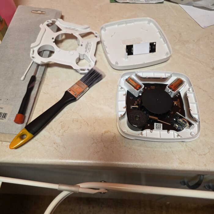 Öppnad rökdetektor med hölje och elektronik synliga, omgiven av rengöringsverktyg som pensel och skruvmejsel på bord.