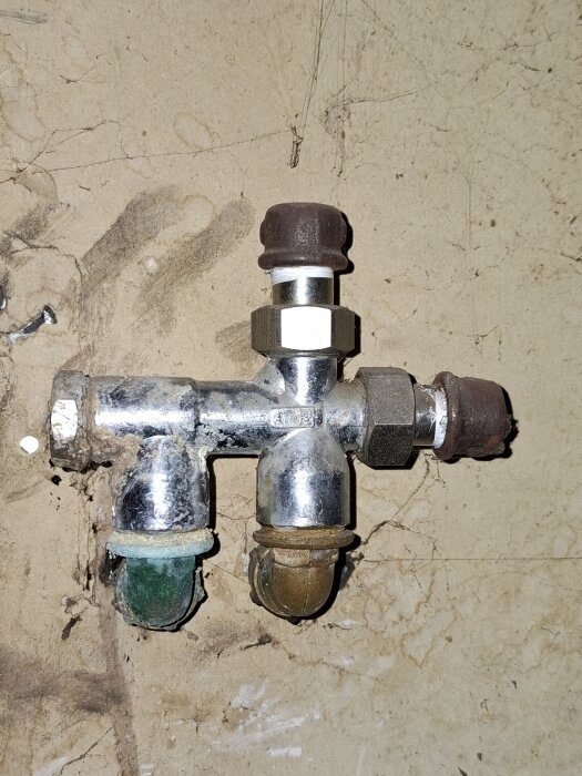 Metallisk VVS-anslutning med tre rör, varav ett grönt och ett brunt, monterad på en vägg med varma rör.