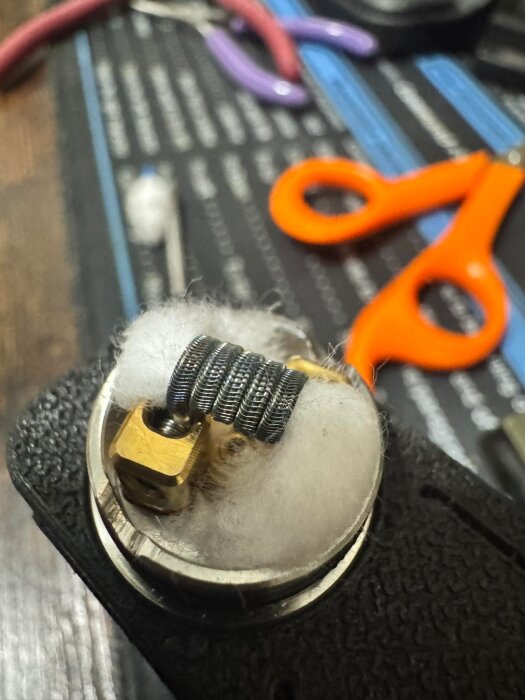 En närbild på en clapton-coil med bomull i en vejp-enhet, orange sax i bakgrunden på ett arbetsverktygsbord.