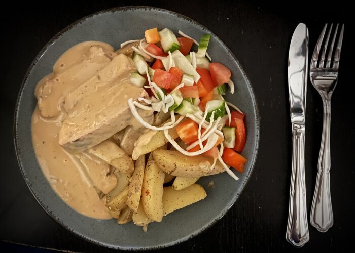 Fläskytterfilé i sås med örtkryddade klyftpotatisar och sallad på tallrik, bredvid bestick på bord.