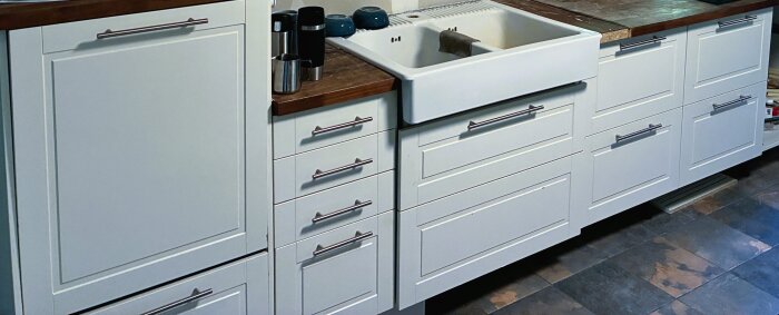 Vitt kök med träbänkskiva, diskho och luckor utan fasad kant. Möjligtvis Marbodal utan bekräftat modellnamn.