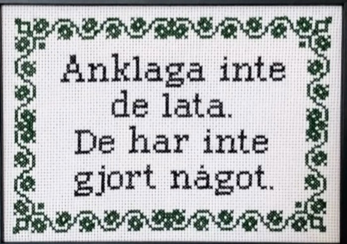 Broderi med texten "Anklaga inte de lata. De har inte gjort något." omgiven av en dekorativ grön bård med blommönster.