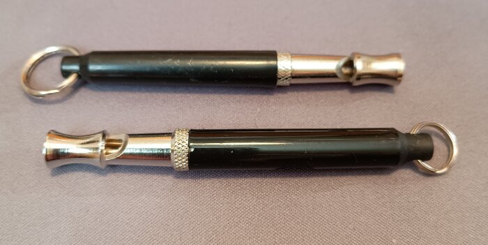 Två svarta hundvisselpipor med silverdetaljer och nyckelringar ligger på en grå yta.