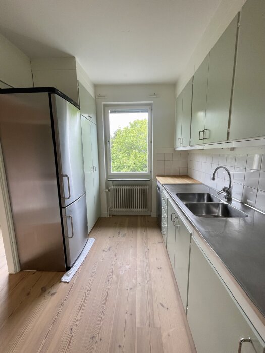 Kök med ljusgröna skåpluckor, rostfri kyl och frys, diskho i rostfritt stål, träbänkskiva och trägolv. Fönster med utsikt över träd.