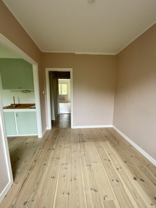 Ljust rum med trägolv, beige väggar, öppning mot kök med gröna skåpluckor och utsikt genom hallen mot ett fönster.