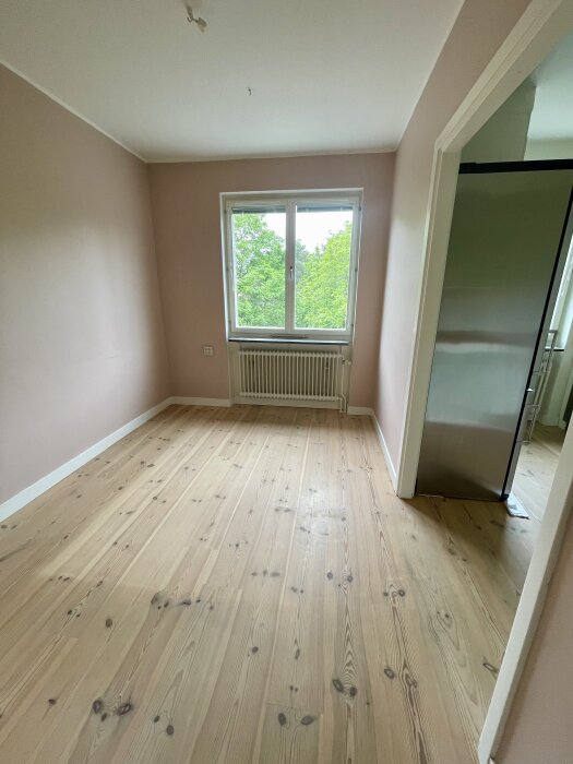 Ett ljust rum med trägolv och ljusrosa väggar, ett fönster med grön utsikt, sett från en dörröppning.