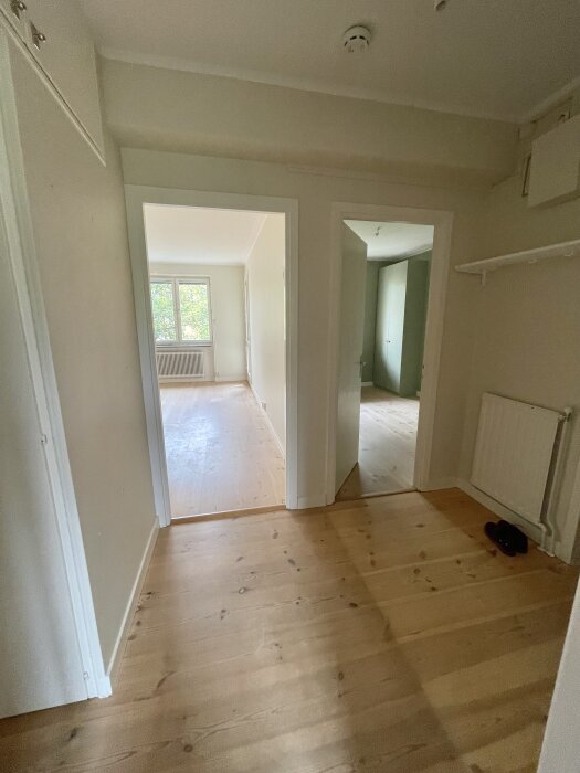 En ljus hall med trägolv och vita dörrkarmar, leder till två rum med fönster och två rum med olika väggfärger. Skor vid elementet.
