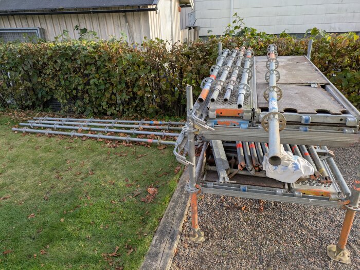 Rullställning delvis ihopmonterad och staplad på en grusplan, med gräs och buskar i bakgrunden, vid ett garage.