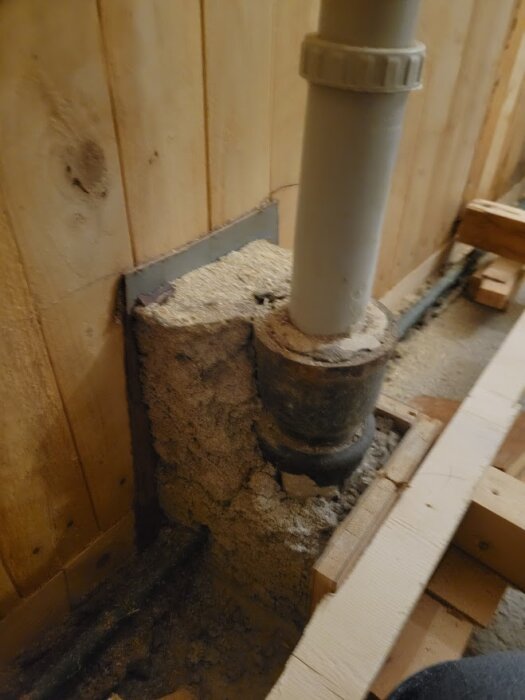 Avlopp under köksdiskbänken med ett rör av gjutjärn, omgivet av betong, del av ett renoveringsprojekt. Träpanel syns i bakgrunden.