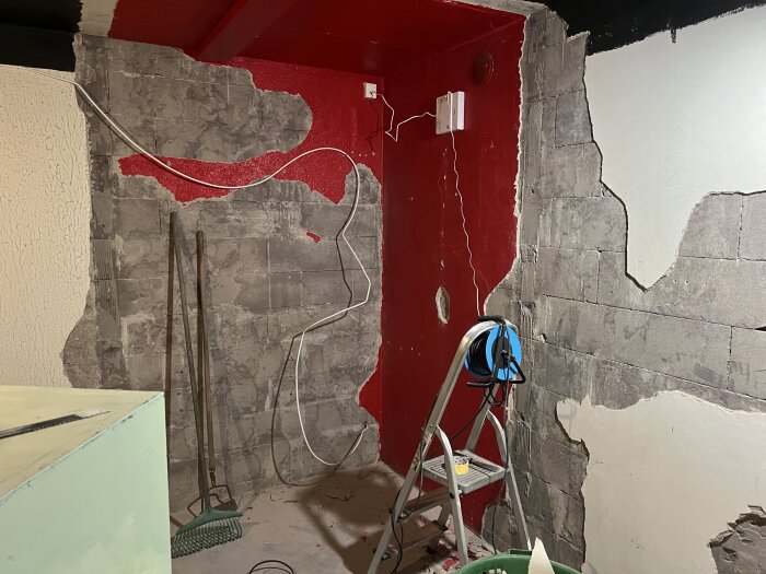 Vägg med borttagen puts visar ojämn yta, stege och verktyg i källarrum. Röd och vit färg syns, elektriska ledningar hänger löst.