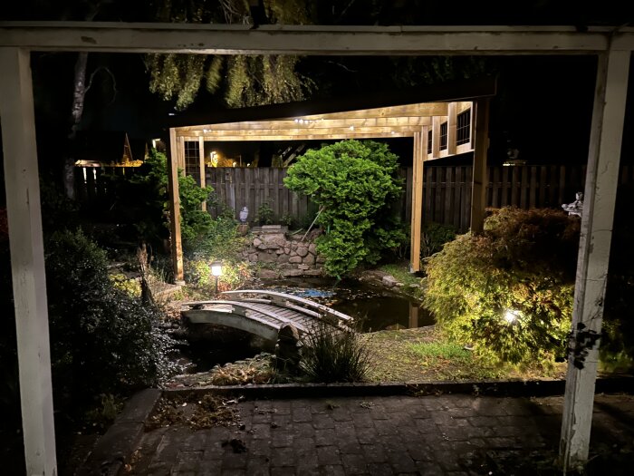 Upplyst trädgård på natten med en liten bro över en damm och en pergola i bakgrunden, omgiven av träd och växter.