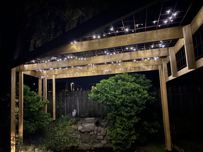Pergola med strålande belysning ovanför en trädgård vid natten, omgiven av grönska och staket i bakgrunden.