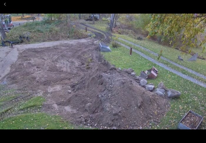 En gräsmatta med grävd jord och stenhögar, troligen från ett pågående avloppsprojekt. Grusväg och träd i bakgrunden.