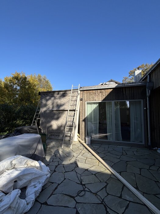 Stege mot husfasad med nyligen monterad panel, takpapp och solcellsdriven markis syns. Byggmaterial och presenningar på stenlagd uteplats.
